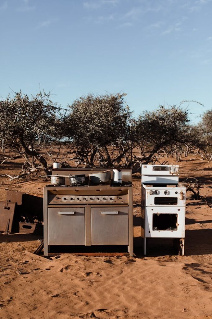 Aged cabinet with kitchenware near stove with oven on sandy land against trees under blue cloudy sky